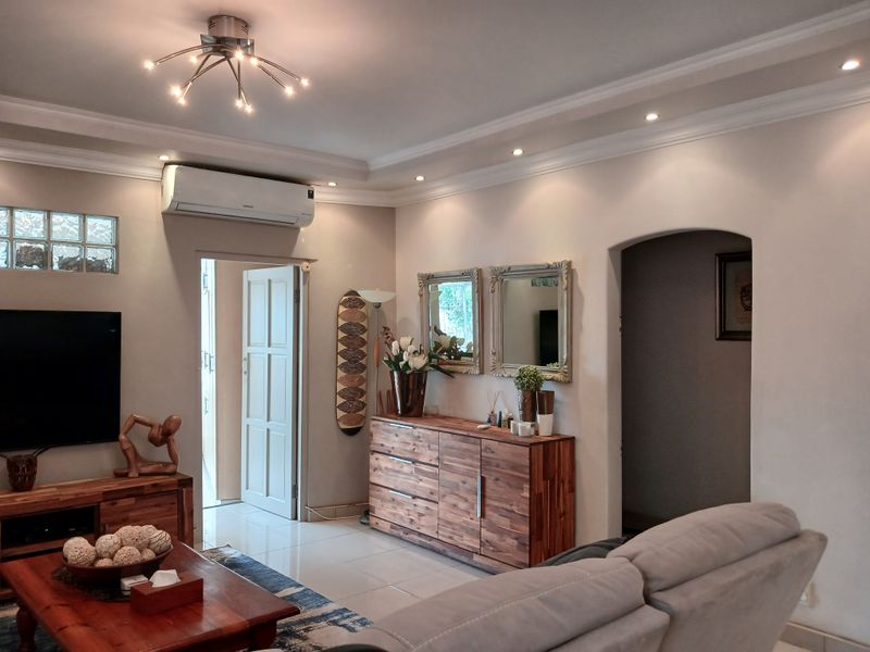 A well-maintained living room featuring tiled flooring, coved cornicing, recessed downlights, and a wall-mounted air conditioning unit.