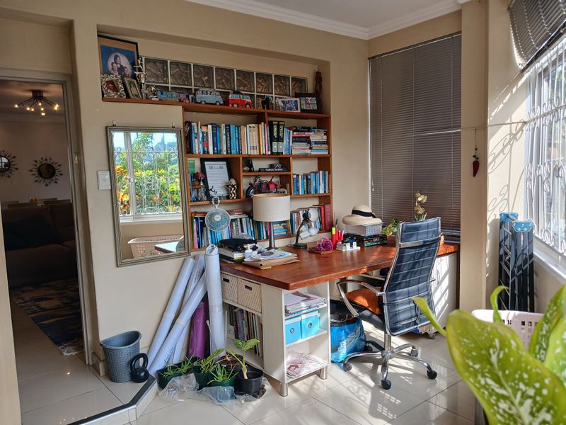 A functional home office space featuring built-in shelving, a glass block accent wall, and large windows with venetian blinds, providing ample natural light.
