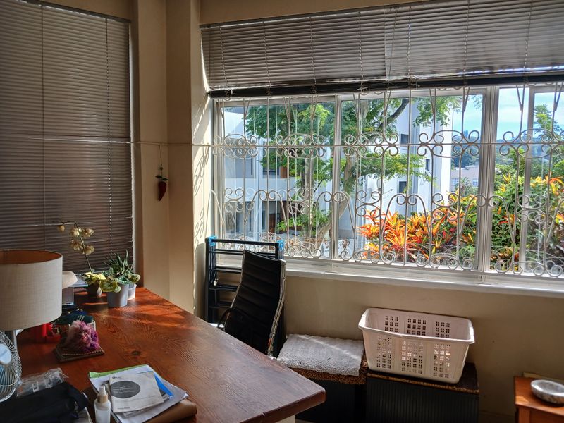 A sunlit office space featuring large sliding windows with decorative wrought iron security bars and adjustable aluminium venetian blinds.