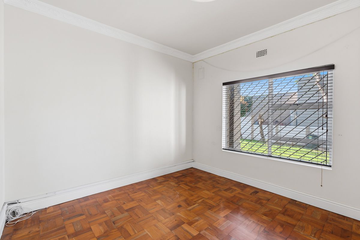 A bright, empty bedroom featuring classic parquet timber flooring, decorative ceiling cornices, and a window fitted with security bars and blinds.