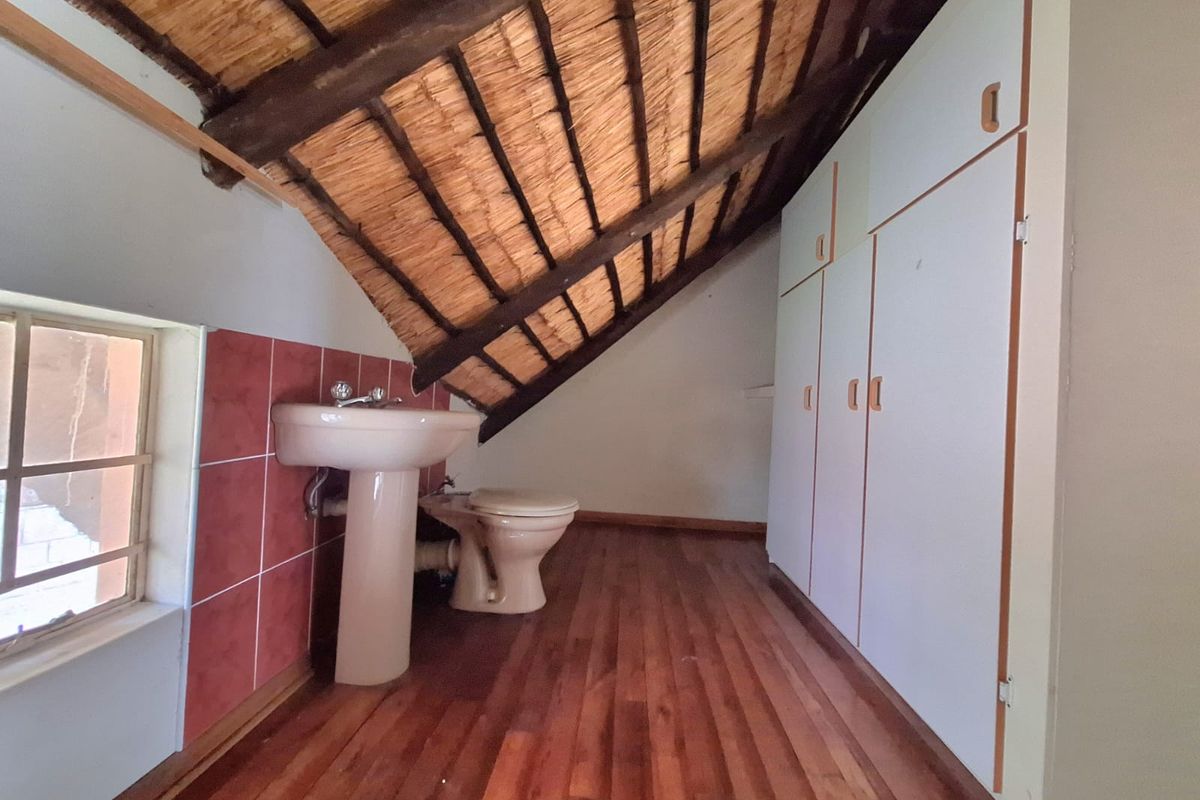 A compact bathroom featuring a pedestal basin, toilet, and built-in storage cupboards under a pitched thatched roof with timber beams.
