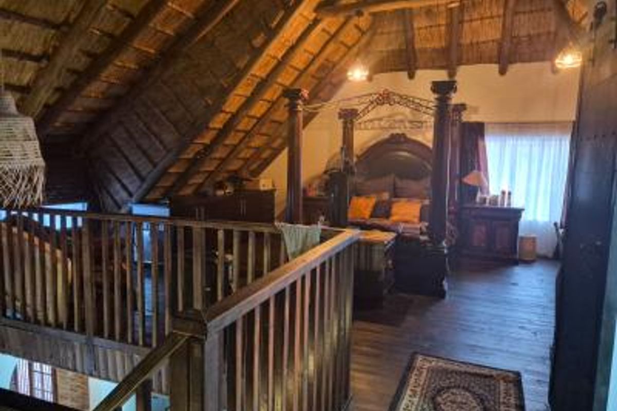 A spacious loft-style bedroom featuring a high thatched roof with exposed timber rafters, wooden flooring, and a mezzanine-level layout.