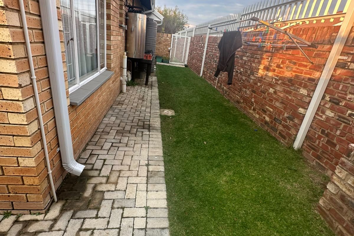 A side exterior view of the townhouse featuring a paved walkway, a small grass lawn, and a brick boundary wall with security spikes.