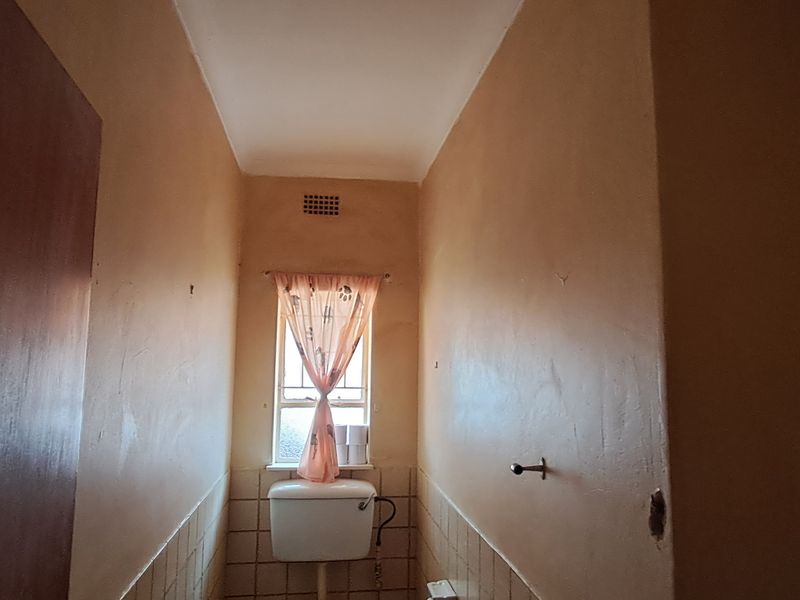 A small, dated bathroom featuring a toilet, ceramic wall tiling, and a window, currently in need of renovation and cosmetic updates.