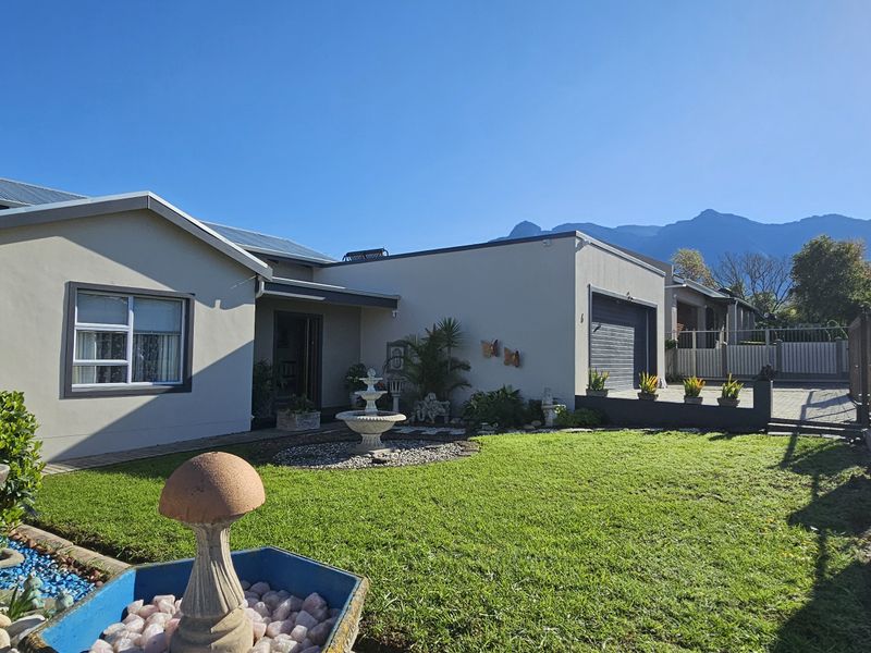 Front view of a well-maintained single-story house featuring a manicured lawn, paved driveway, garage, and secure boundary walling against a mountain backdrop.