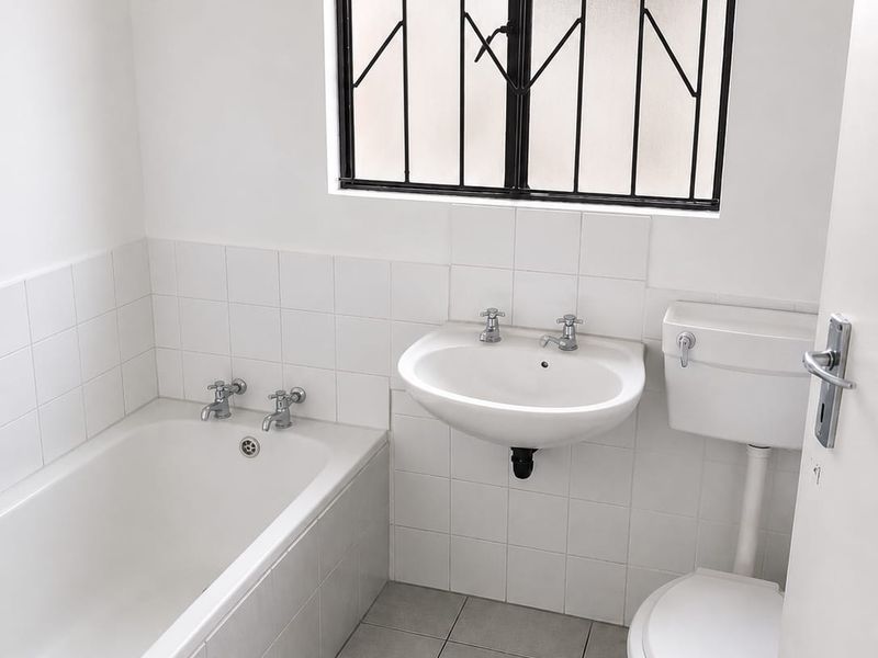 A clean, functional bathroom featuring a white bathtub, wall-mounted basin, and toilet with neutral tiled finishes and security bars on the window.