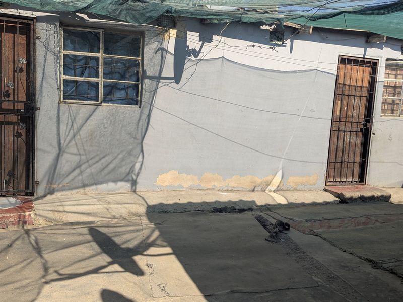 Rear exterior view of the property featuring concrete paving, security gates on doors and windows, and overhead shade netting.