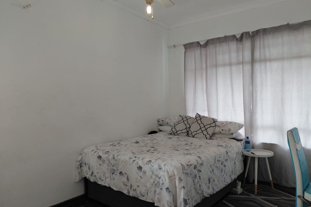 A bedroom featuring parquet flooring, a ceiling fan, and large windows with curtain rails, offering a functional space ready for cosmetic updates.