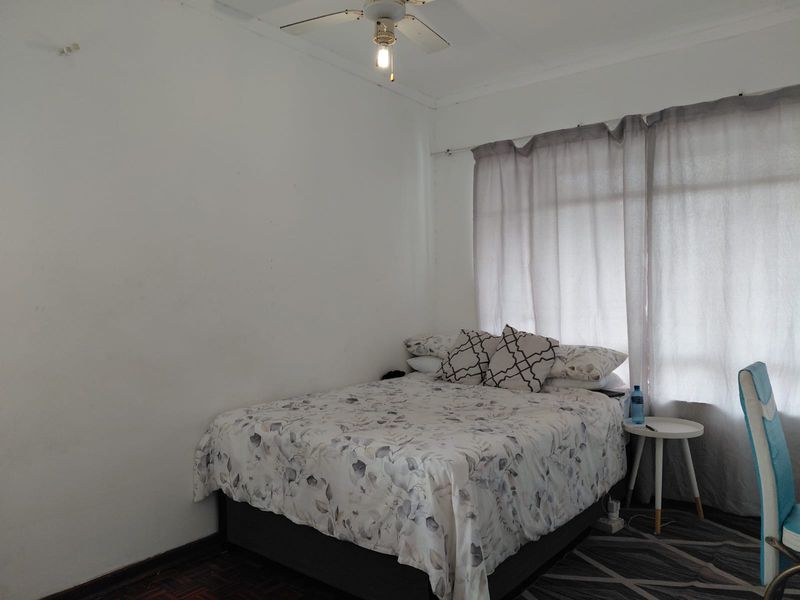 A bedroom featuring parquet flooring, a ceiling fan, and large windows with curtain rails, offering a functional space ready for cosmetic updates.
