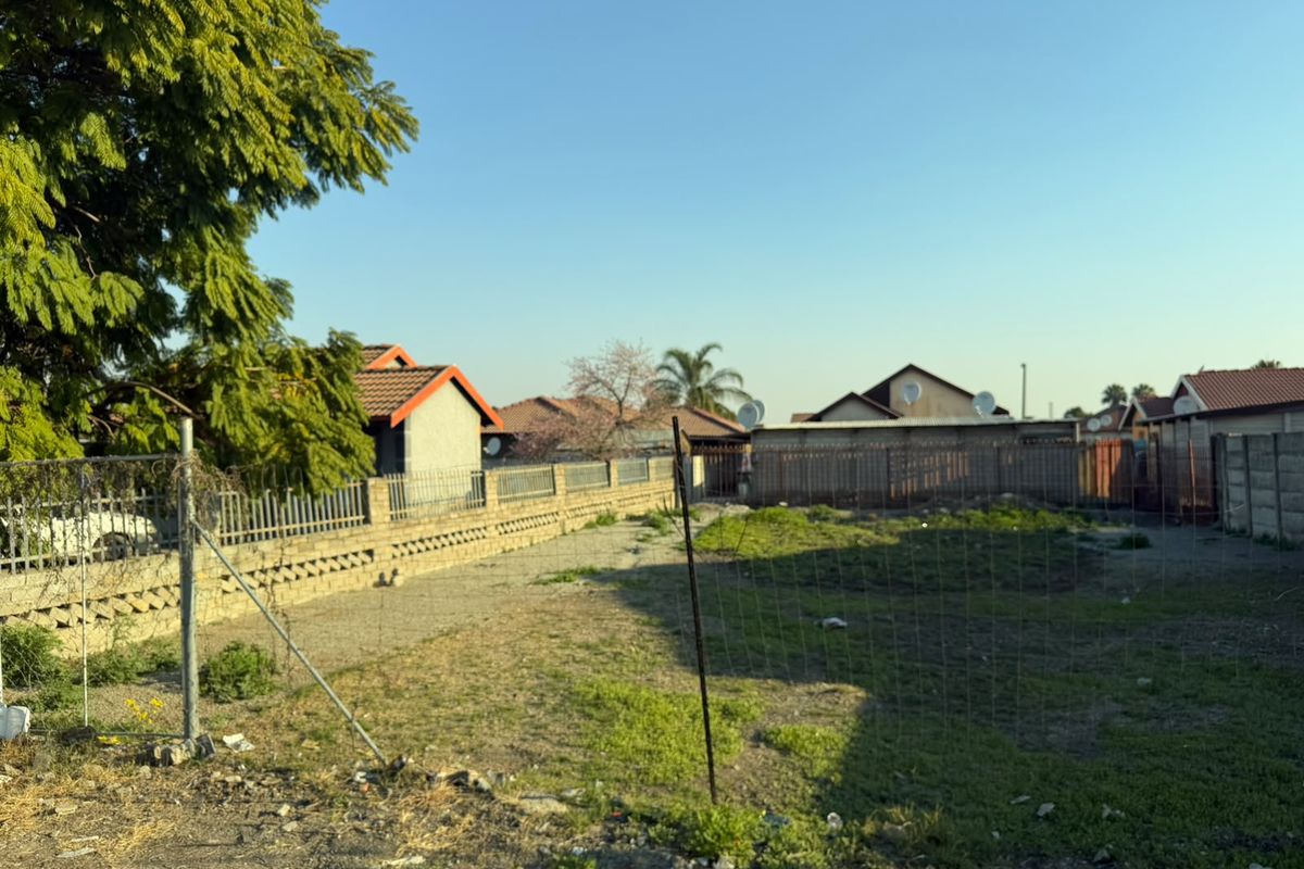A vacant residential plot featuring perimeter walling, wire mesh fencing, and a metal gate, offering a clear space for potential development.