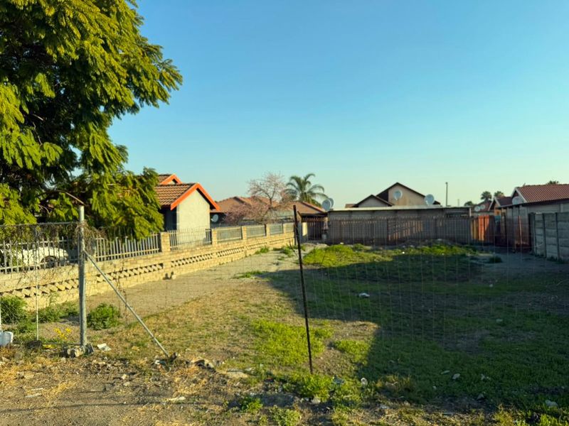 A vacant residential plot enclosed by perimeter fencing and boundary walls, featuring natural ground cover and established trees.