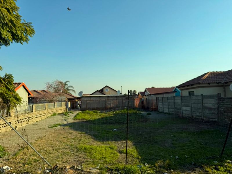 A vacant residential plot enclosed by perimeter walls and wire fencing, featuring an open grass area with some debris.