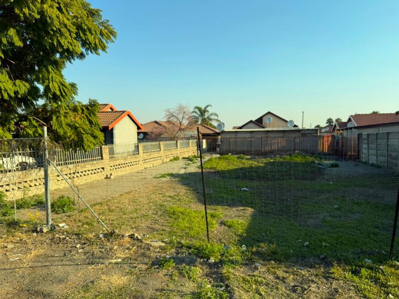 A vacant residential plot featuring perimeter walling and wire mesh fencing, showing signs of overgrown vegetation and surface debris.