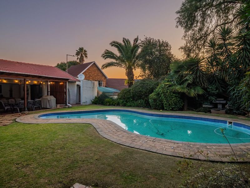 A well-maintained backyard featuring a curved swimming pool with tiled coping, adjacent to a covered patio area and established garden landscaping.
