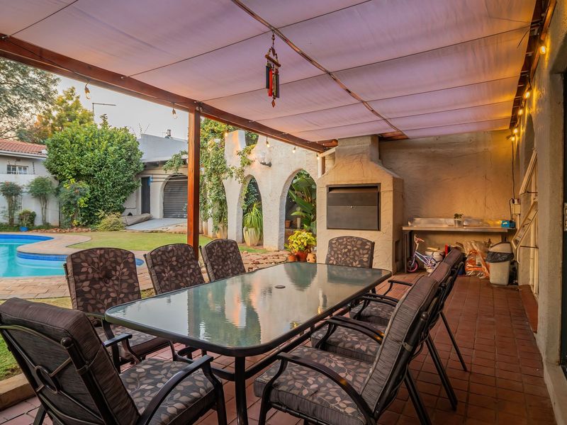 Covered outdoor patio featuring a built-in braai, sink, and tiled flooring, overlooking a private swimming pool and garden area.