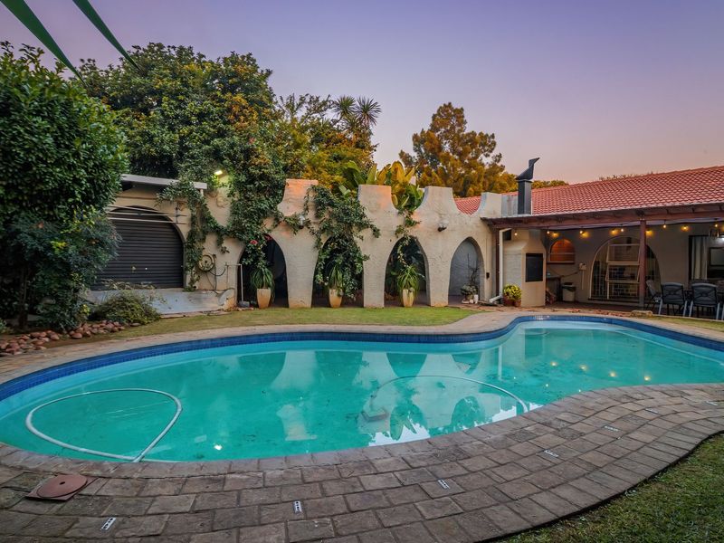 Outdoor pool area featuring a paved deck, covered patio with built-in braai, and unique arched architectural wall elements set within a landscaped garden.