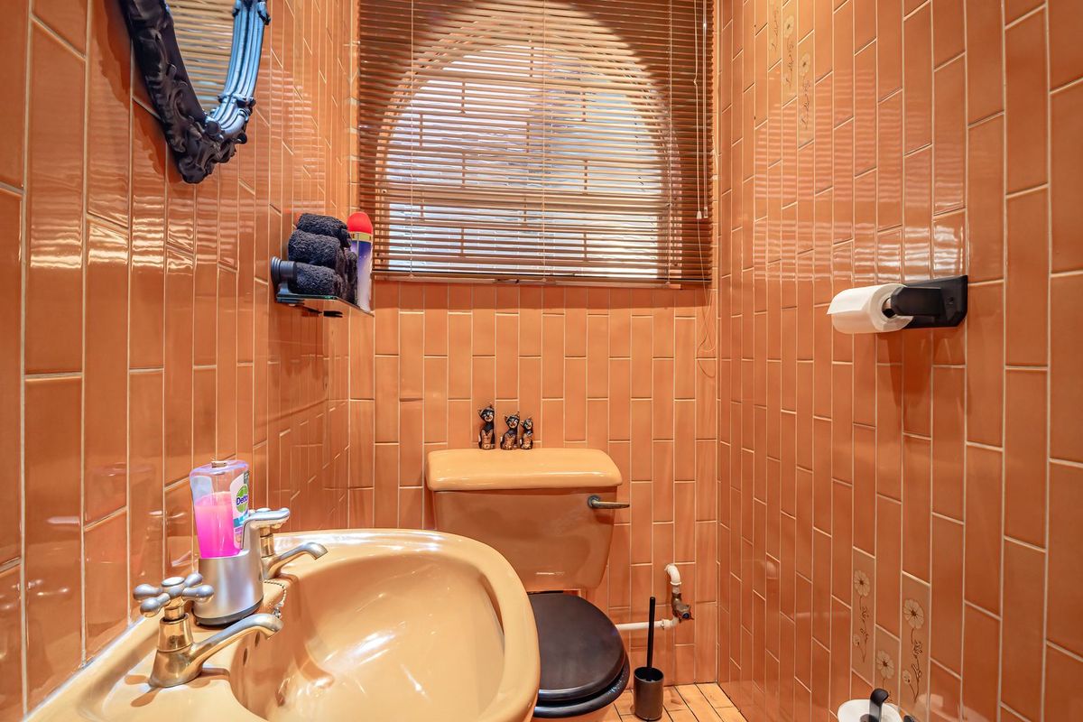A compact bathroom featuring floor-to-ceiling orange ceramic wall tiles, a pedestal basin, and a toilet, illuminated by an arched window with venetian blinds.