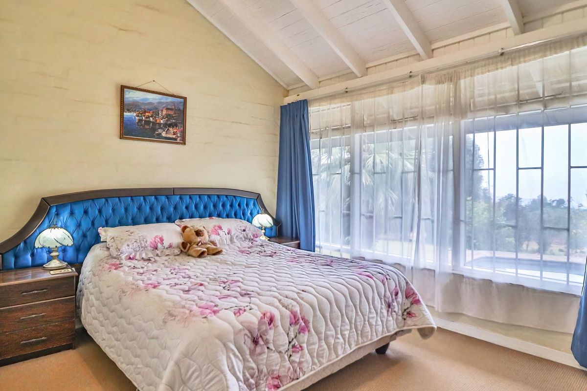A cozy bedroom featuring a vaulted ceiling with exposed timber beams, carpeted flooring, and a large window with security bars allowing natural light.