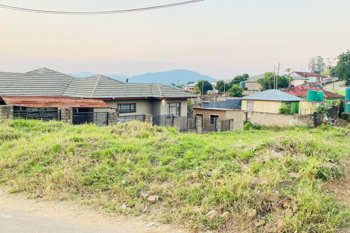 A view of a residential plot featuring uneven, grass-covered terrain with surrounding neighborhood houses and perimeter fencing visible in the background.
