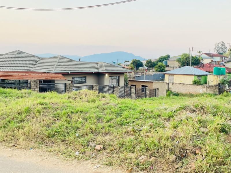 A view of a residential plot featuring uneven, grass-covered terrain with surrounding neighborhood houses and perimeter fencing visible in the background.