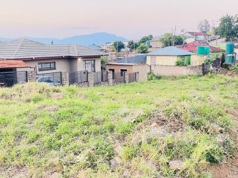 A view of a vacant residential plot featuring grassy terrain, enclosed by boundary walls and fencing, situated within an established neighborhood.