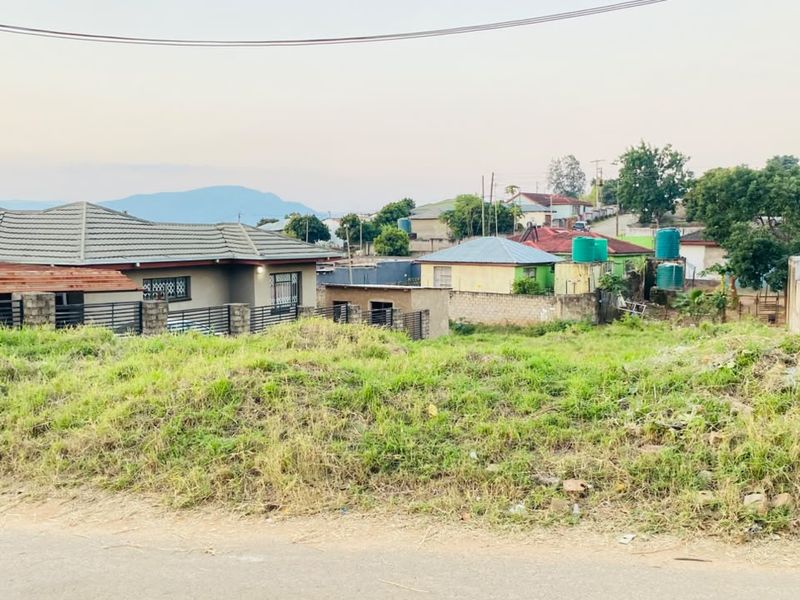 A vacant residential plot featuring natural grass coverage, boundary walls, and views of the surrounding neighborhood and distant mountains.