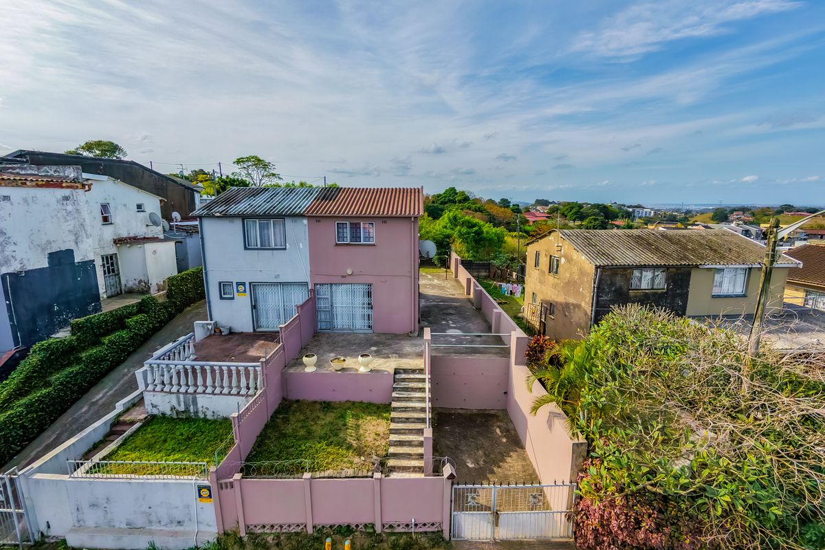 Aerial view of a two-story townhouse property featuring a paved driveway, perimeter wall with security fencing, and a tiered backyard area.