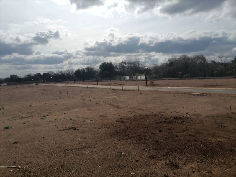 A view of a vacant residential plot featuring a paved road, perimeter fencing, and established street lighting.