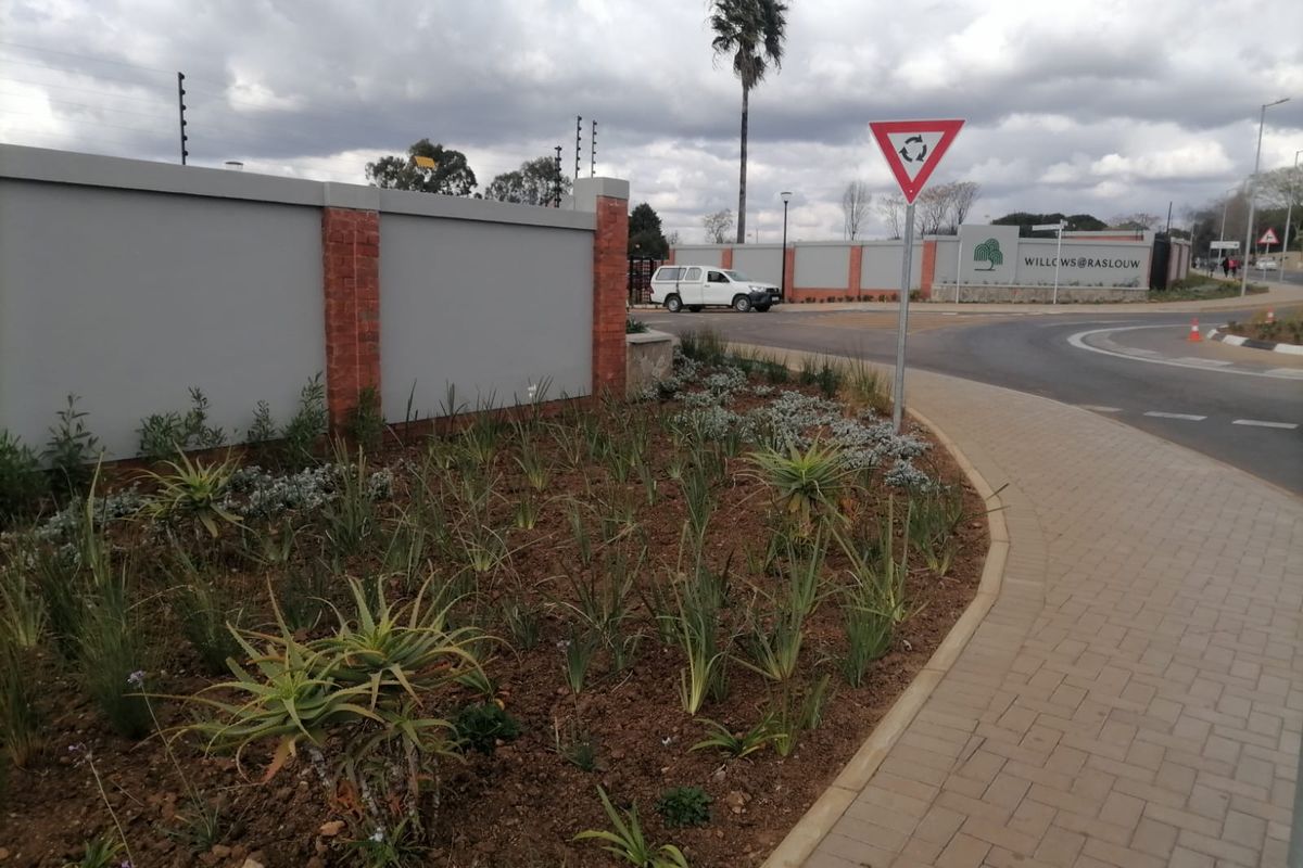 Street view of the estate entrance featuring a perimeter wall with electric fencing, paved sidewalk, and landscaped verge.
