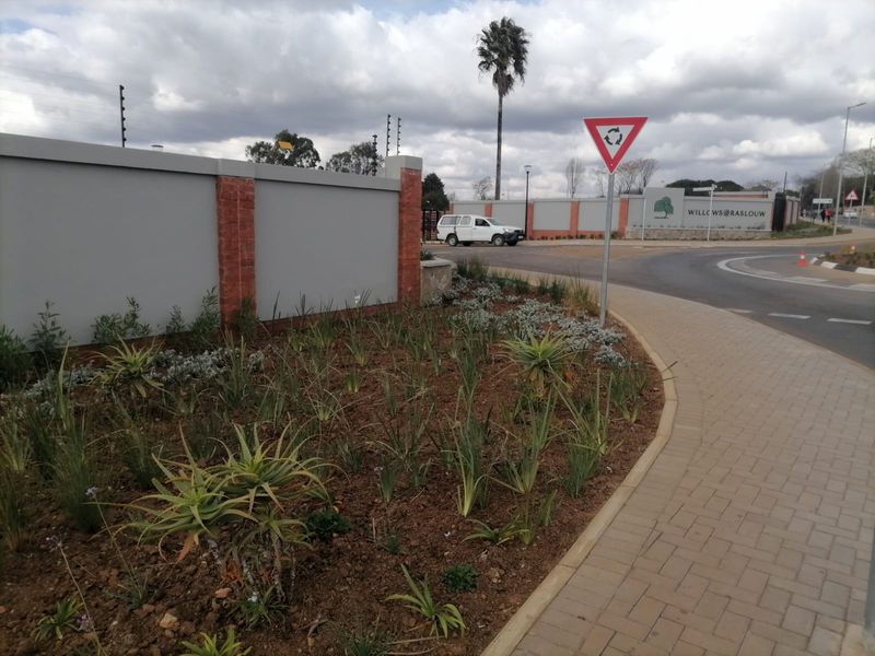Street view of the estate entrance featuring a perimeter wall with electric fencing, paved sidewalk, and landscaped verge.