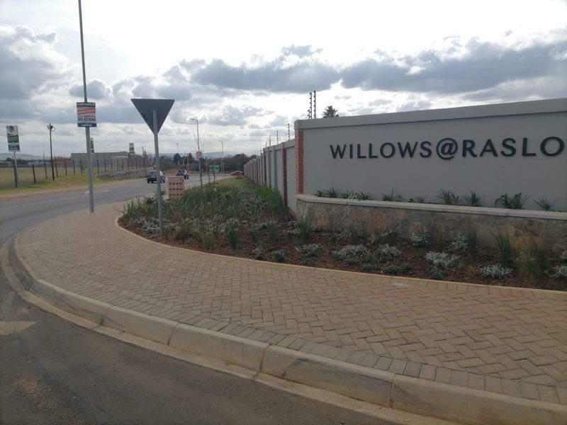 Street view of the Willows at Raslo residential estate entrance, featuring a paved sidewalk, perimeter wall with electric fencing, and landscaped verge.