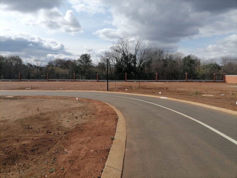 A view of a paved residential street within a vacant land development, featuring concrete curbing and a perimeter brick boundary wall.