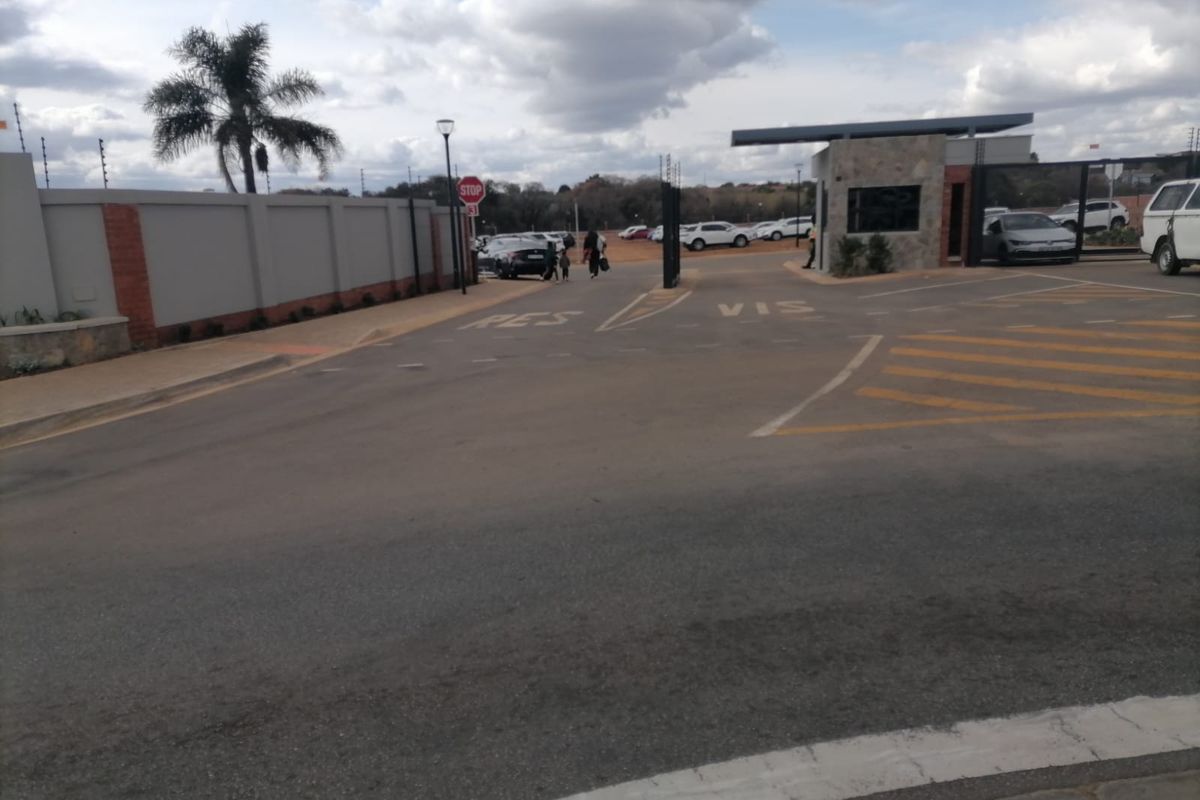 View of the residential estate entrance featuring a security gatehouse, perimeter walling with electric fencing, and paved access roads.