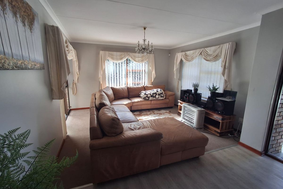 A spacious living room featuring a mix of carpet and laminate flooring, large windows with security bars, and a central chandelier.