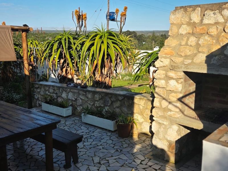 Outdoor patio area featuring a stone-clad built-in braai, crazy paving flooring, and a pergola, overlooking a garden with mature aloe plants.