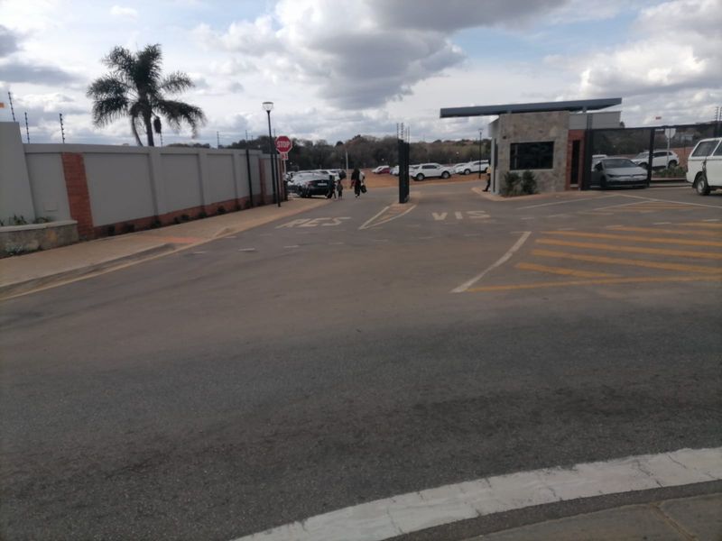 View of the residential estate entrance featuring a security gatehouse, perimeter walling with electric fencing, and paved access roads.
