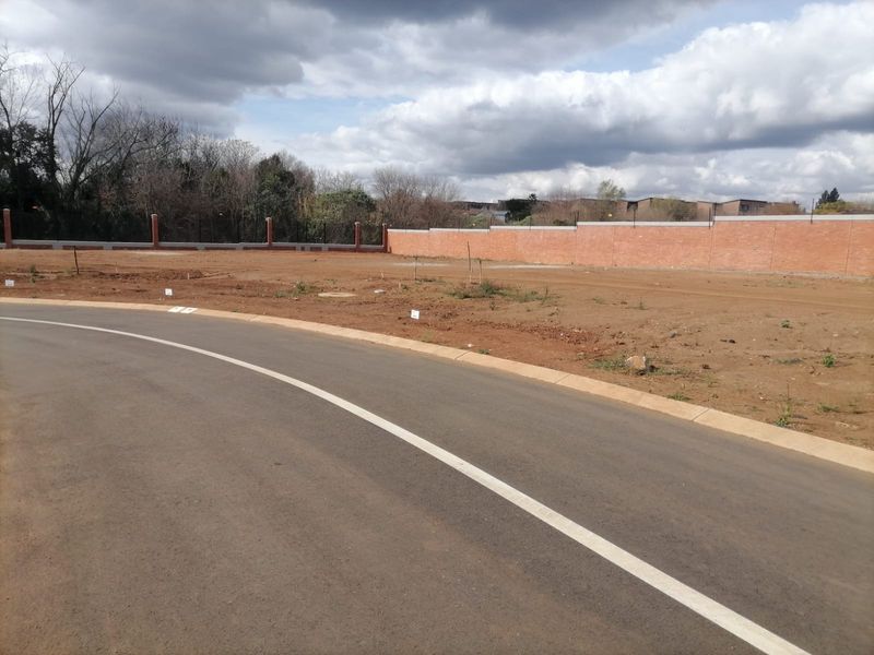 A view of a vacant residential plot featuring a paved road, concrete curbing, and a secure brick boundary wall with electric fencing.