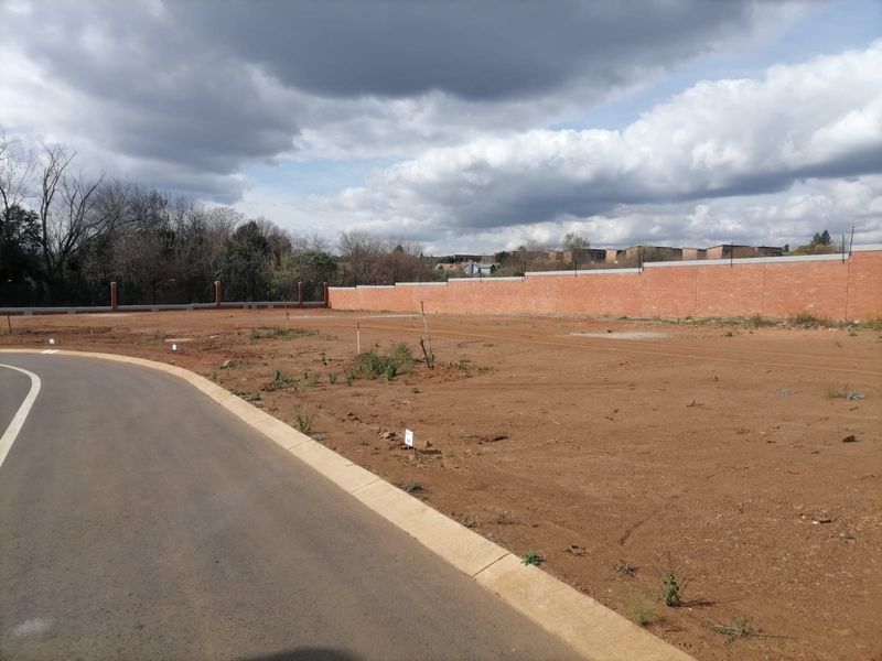 A vacant residential plot featuring a paved road, concrete curbing, and a secure brick perimeter wall with electric fencing.