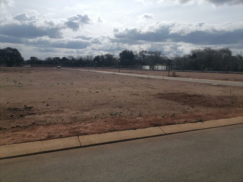 A vacant residential plot featuring a level surface, paved road access, concrete curbing, and perimeter fencing in a developed estate setting.