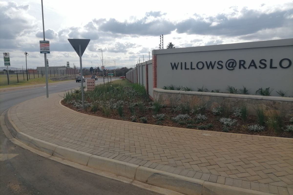 Street view of the Willows at Raslo residential estate entrance, featuring a paved sidewalk, perimeter wall with electric fencing, and landscaped verge.