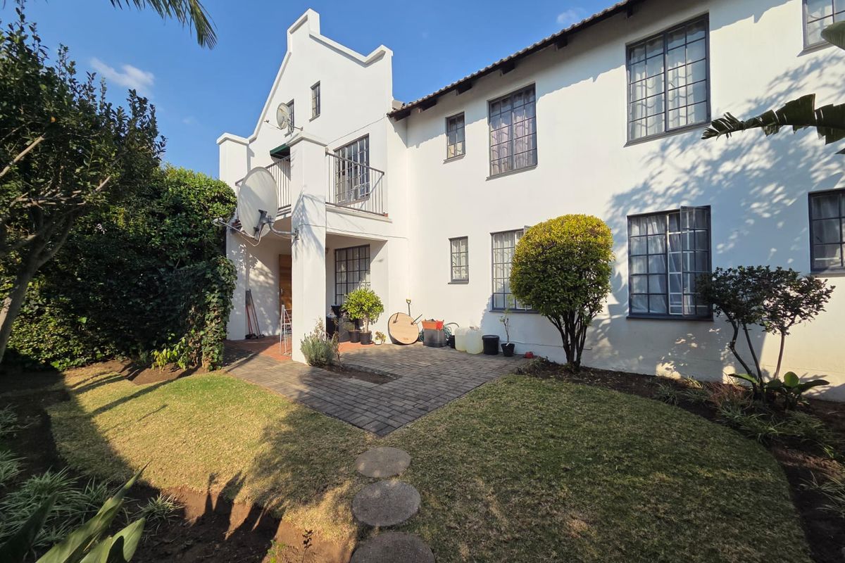 Rear view of a townhouse featuring a paved patio area, a small balcony, and well-maintained garden space with gabled architectural detailing.