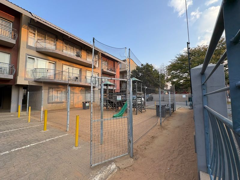 Exterior view of an apartment complex featuring a fenced playground area, paved parking, and multiple balconies with sliding door access.