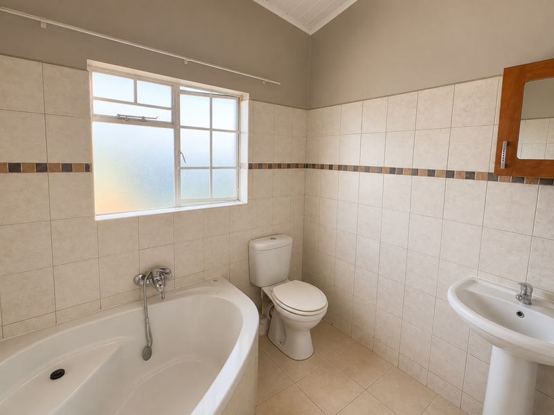 A clean, functional bathroom featuring a corner bathtub, pedestal basin, and toilet, finished with neutral wall and floor tiling.