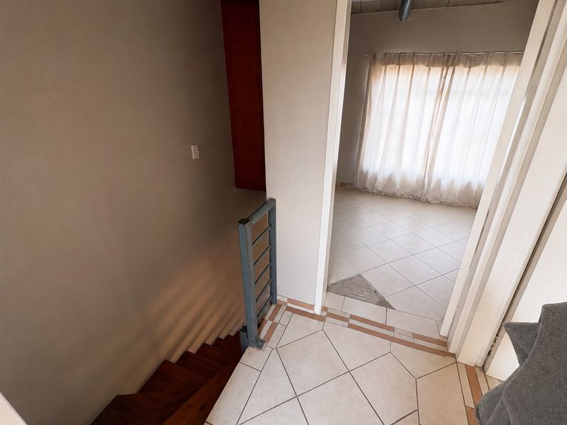 A hallway area featuring a wooden staircase with a metal balustrade, leading into an adjacent room with tiled flooring and large window curtains.