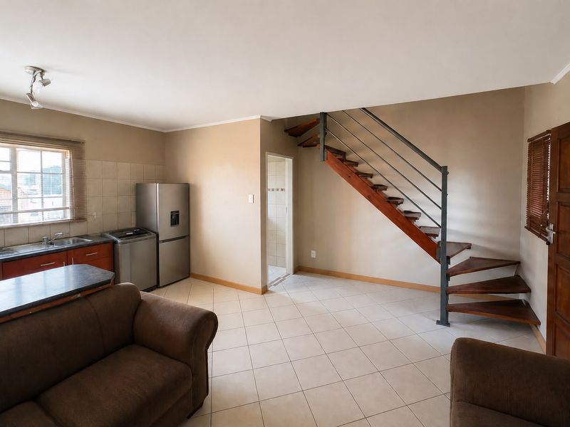 Open plan living area featuring tiled flooring, a wooden staircase with metal railing, and a functional kitchen corner with sink and tiled backsplash.
