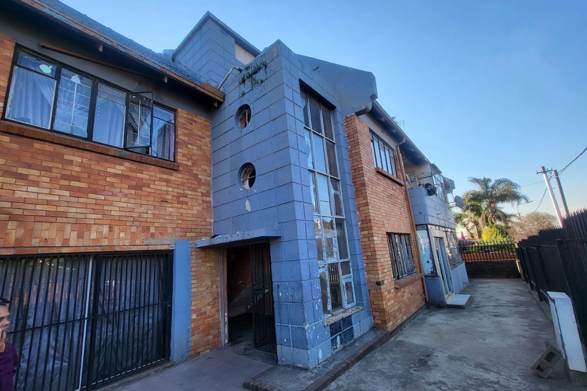 Rear exterior view of a multi-story apartment building featuring brick and painted masonry facade with security bars and concrete ground area.