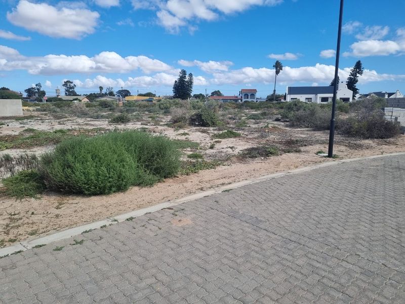 A vacant residential plot of land viewed from the adjacent paved street, featuring natural vegetation and surrounding neighborhood context.