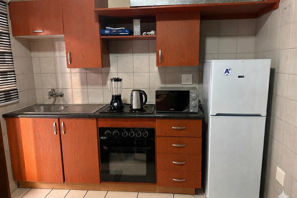 Compact kitchen featuring wooden cabinetry, built-in oven, electric hob, stainless steel sink, and tiled backsplash with neutral floor tiles.