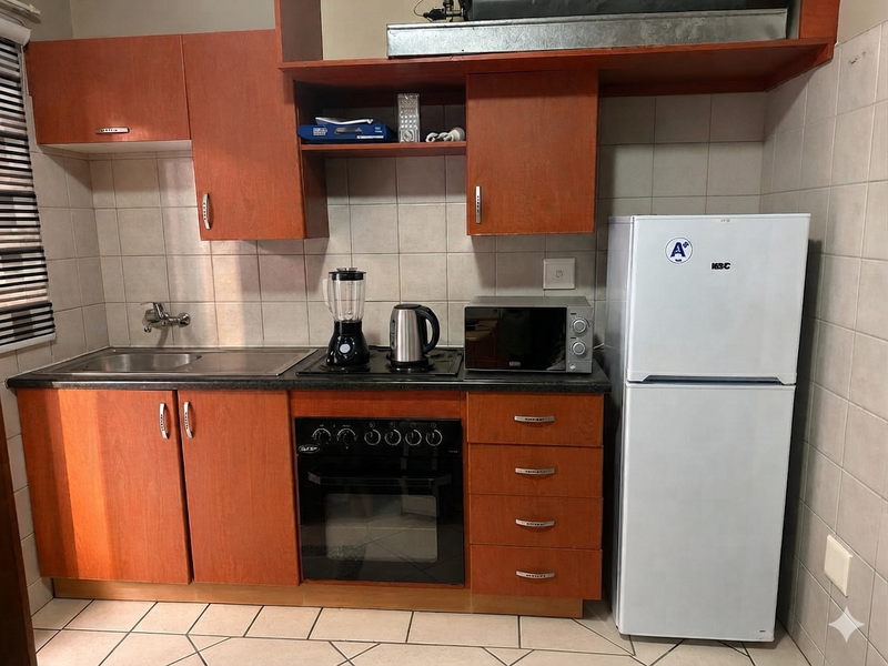 Compact kitchen featuring wooden cabinetry, built-in oven, electric hob, stainless steel sink, and tiled backsplash with neutral floor tiles.