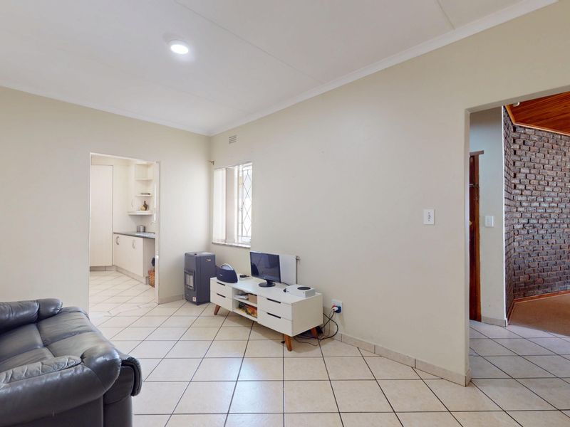 A living room featuring tiled flooring and recessed lighting, with an open-plan flow leading to a kitchen area and an adjacent room with brick walling.